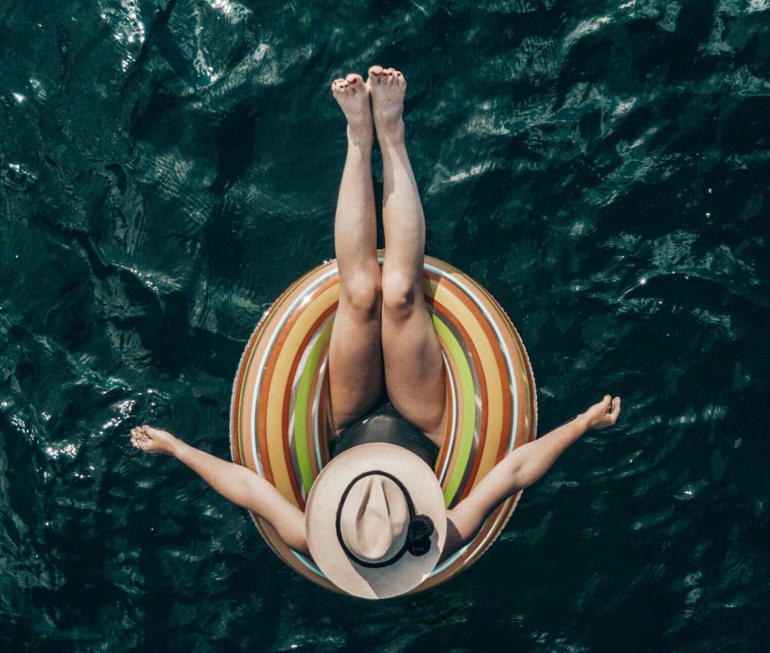 Top view of unrecognizable barefoot tourist in hat lying in inflatable ring on rippled sea during summer trip